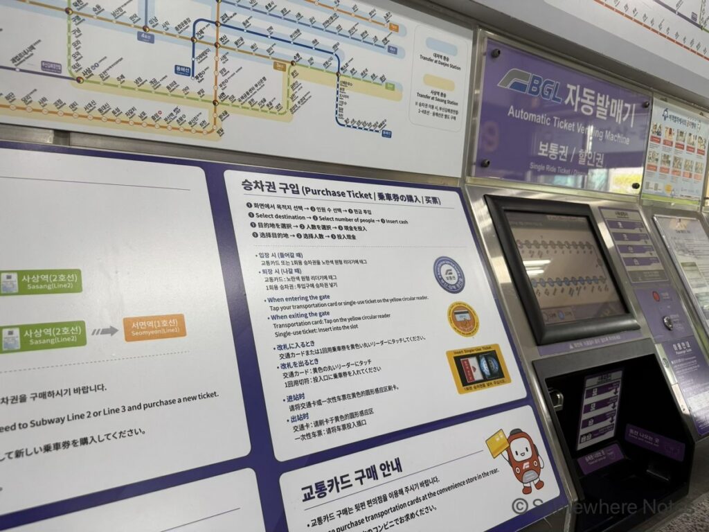 釜山金海軽電鉄の自動券売機の写真。タッチパネル画面の上には紫色の「BGL Automatic Ticket Vending Machine」の看板がある。画面下には大きな多言語案内パネルがあり、「乗車券の購入」という日本語を含む見出しと、日本語を含む手順説明（行先・人数・現金）、および改札での使い方（日本語対応）が、イラストとともに詳しく掲載されている。券売機の上部には路線図があり、下部にはお釣り受け取り口も見える。