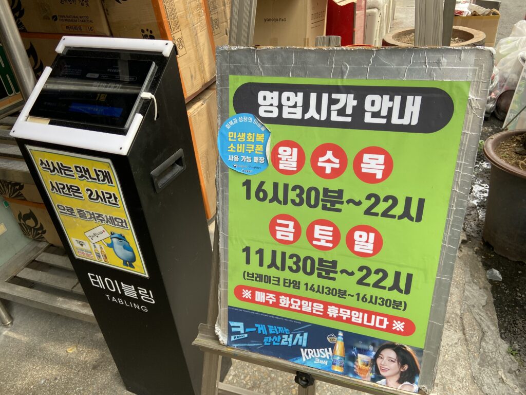 永登浦のプイルスップルカルビの営業時間と定休日が書かれた看板。横にはタッチパネル式の番号札発券機がある。