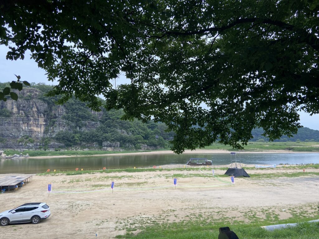 川の向こうにそびえる、岩山の絶景スポット「芙蓉台（プヨンデ）」。手前の川岸には砂地が広がり、緑豊かな木々が見える。
