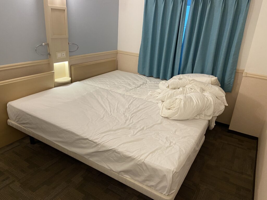 東横インヨンドゥンポの客室にあるツインの部屋。子どもがベッドから落ちないようにくっつけている。白い布団カバーがかかっており、ヘッドボードの脇には間接照明と棚、水色のカーテンが見える。