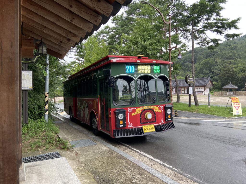 A red and green trolley bus at a stop in Hahoe Folk Village, heading toward Andong Station. Behind the bus is a small ticket booth, surrounded by trees and a parking area.
