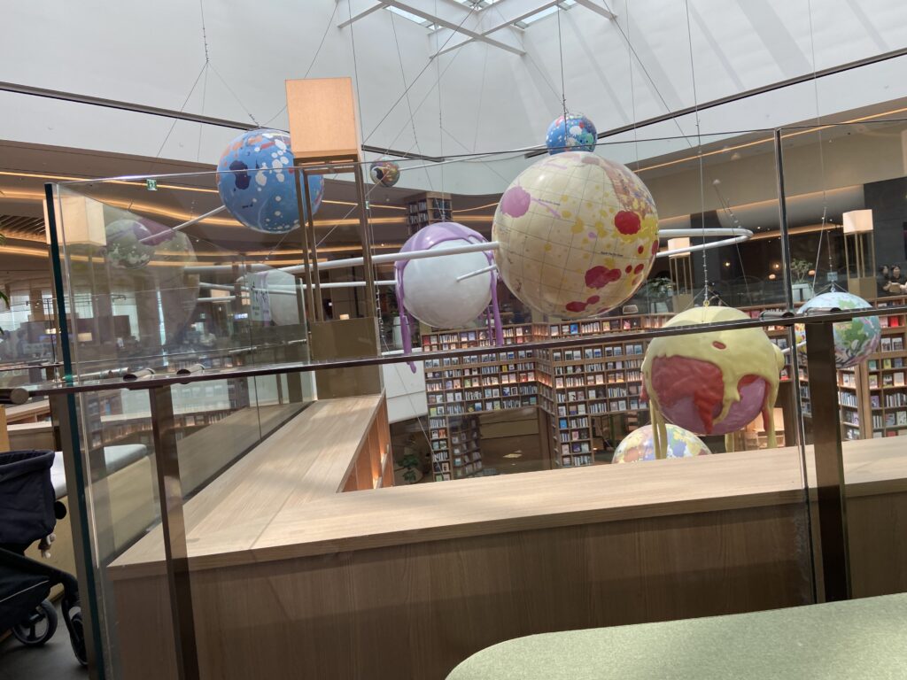 スターフィールドスウォンのピョルマダン図書館にある席からの視点。中央には地球儀を模したカラフルなアート作品が吊るされ、その奥には何層にも重なる巨大な本棚が見える。手前の席には黄緑色のベンチ型クッションの椅子が配置され、人々が休憩している開放的な空間。