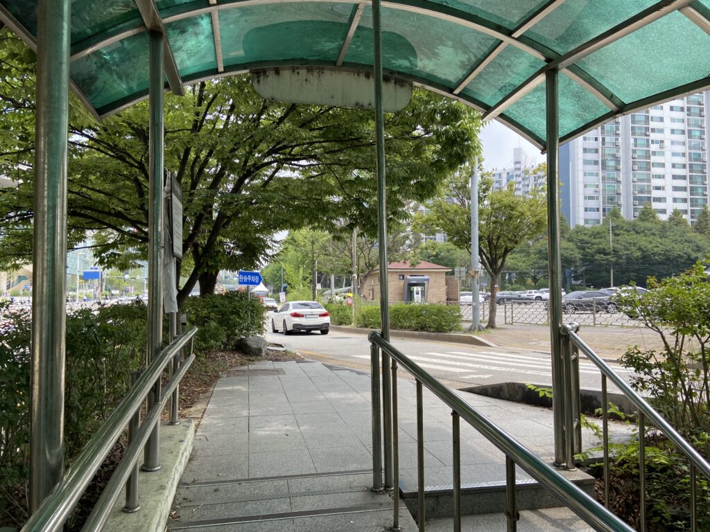 地下鉄ファソ駅の出口から地上に出る階段を上がり、通路を抜けた先に横断歩道と車道が見え、奥にはアパートなどの建物が並んでいる。