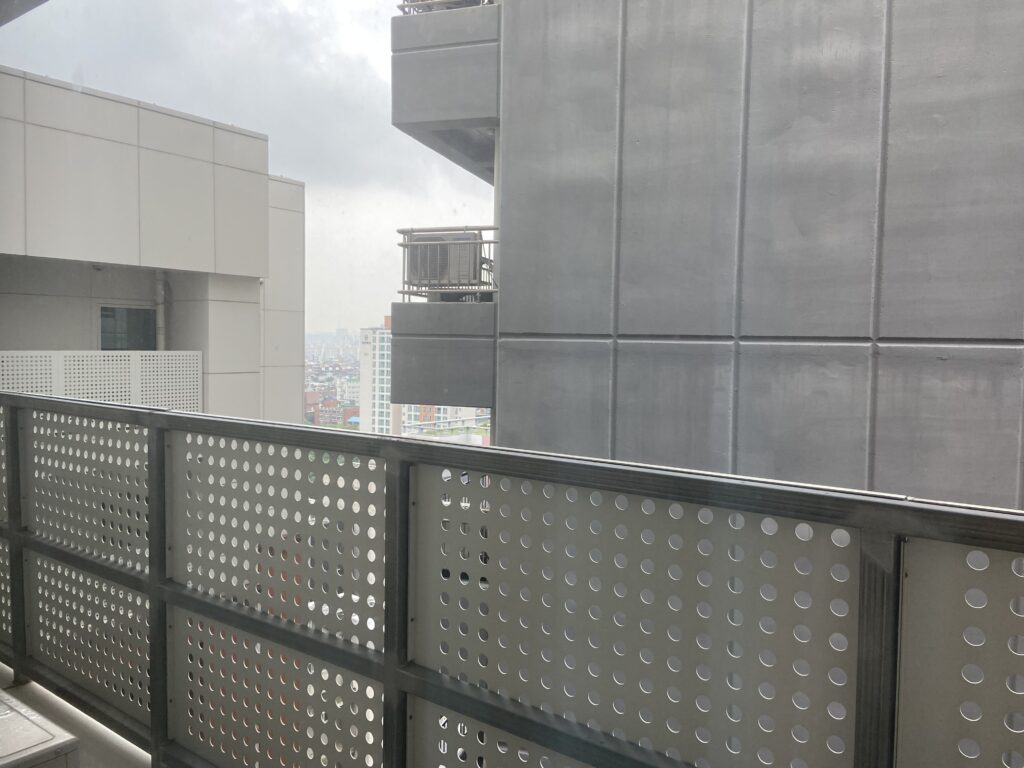 高層ビルとアパートのような現代的な建物が並ぶ風景。手前には丸い穴の開いた金属製のフェンスがある。