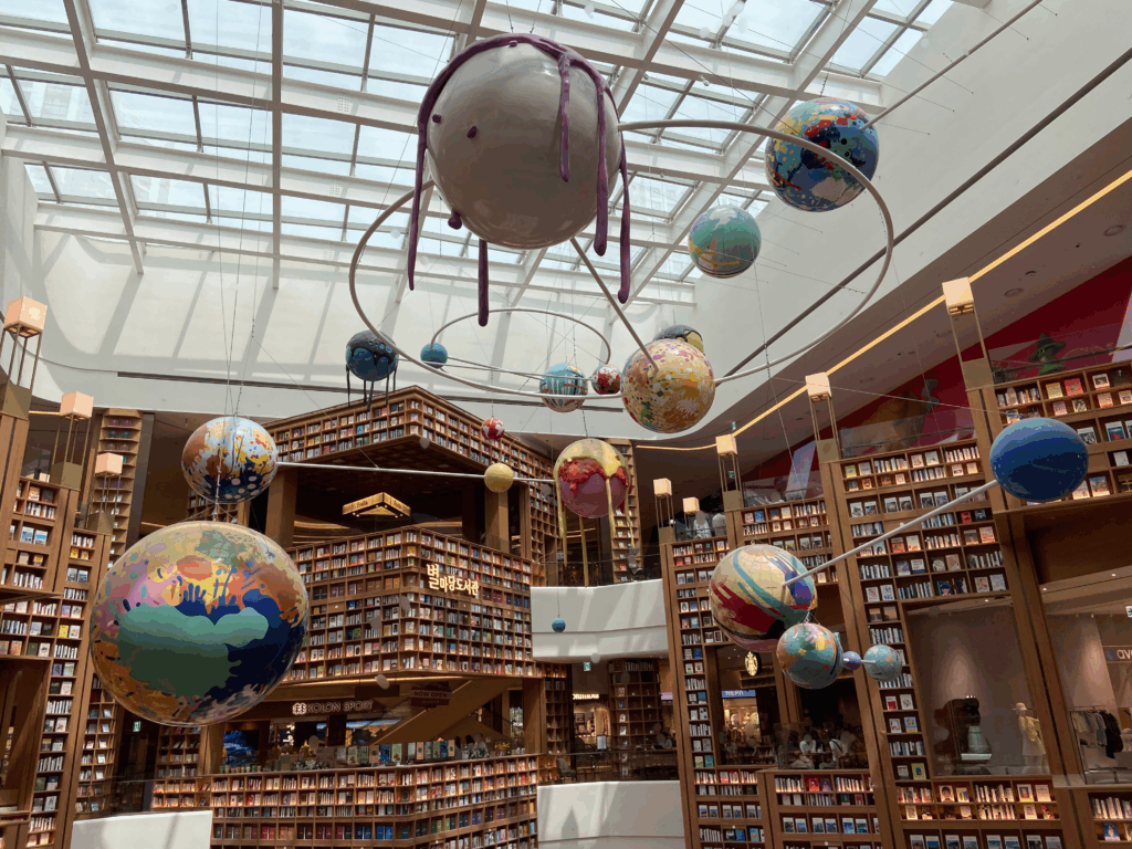 ピョルマダン図書館のアート。スターフィールドスウォンのピョルマダン図書館の巨大な吹き抜け空間。天井から様々なデザインの地球儀をモチーフにしたアート作品が吊るされており、奥には何層にも重なった木製の本棚が見える。