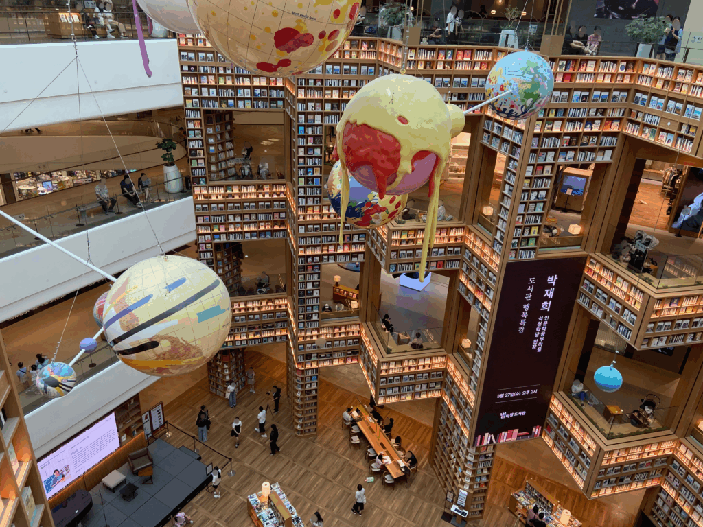 スターフィールド水原のピョルマダン図書館の壮大な全景。何階層にもわたる巨大な本棚の間に、地球儀をモチーフとしたカラフルなアート作品が吊るされ、人々が本棚のテラス席や中央のテーブルでくつろいでいる様子。韓国旅行の休憩や撮影に最適な、アートのような空間。
