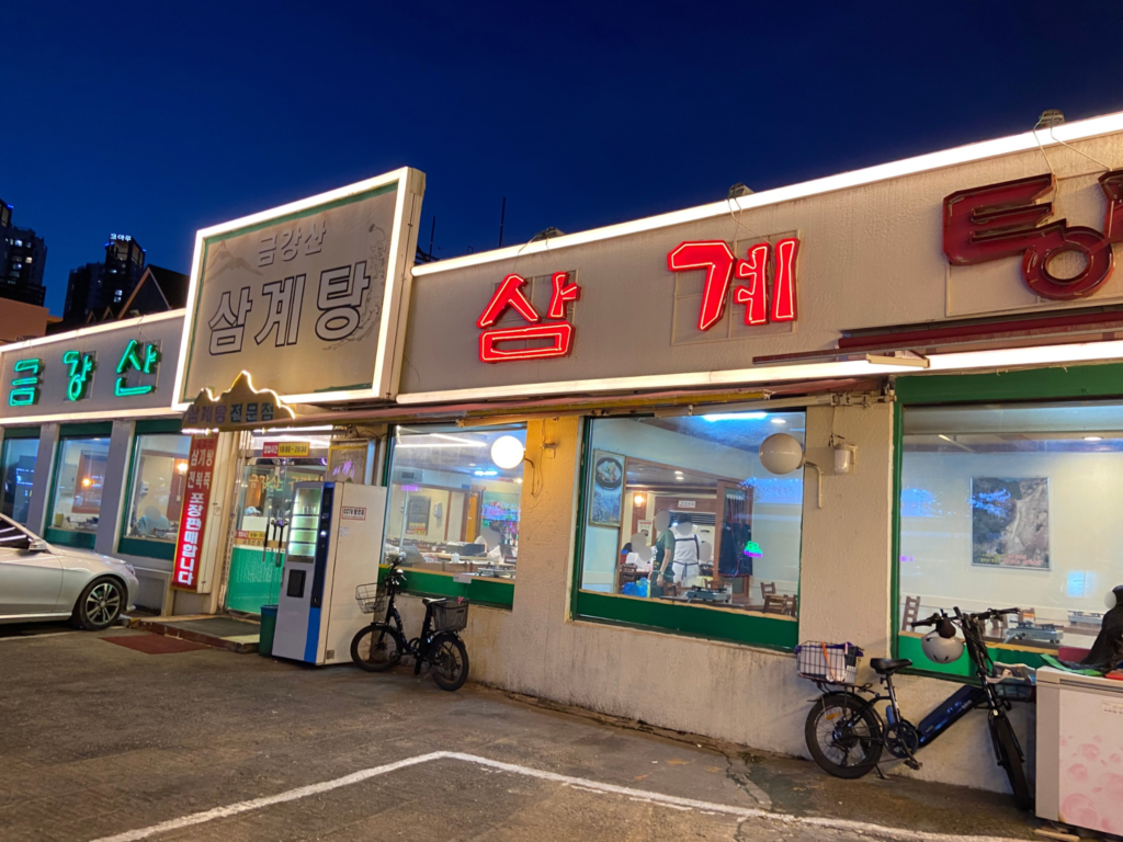 夜に撮影したクムガンサンサムゲタンの店舗外観。大きく『サムゲタン』と書かれた看板や、赤と緑のネオンサインが明るく点灯している。
