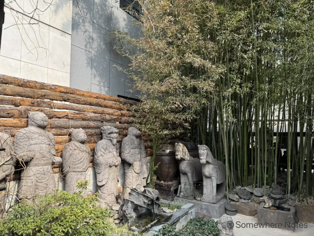 釜山のモモスコーヒー本店の入り口入ってすぐの場所。木のログフェンスの前に、人型、動物型などの石像が並んでいる。右側には、青々とした背の高い緑の竹林が茂り、その奥にカフェのテラス席が見える。