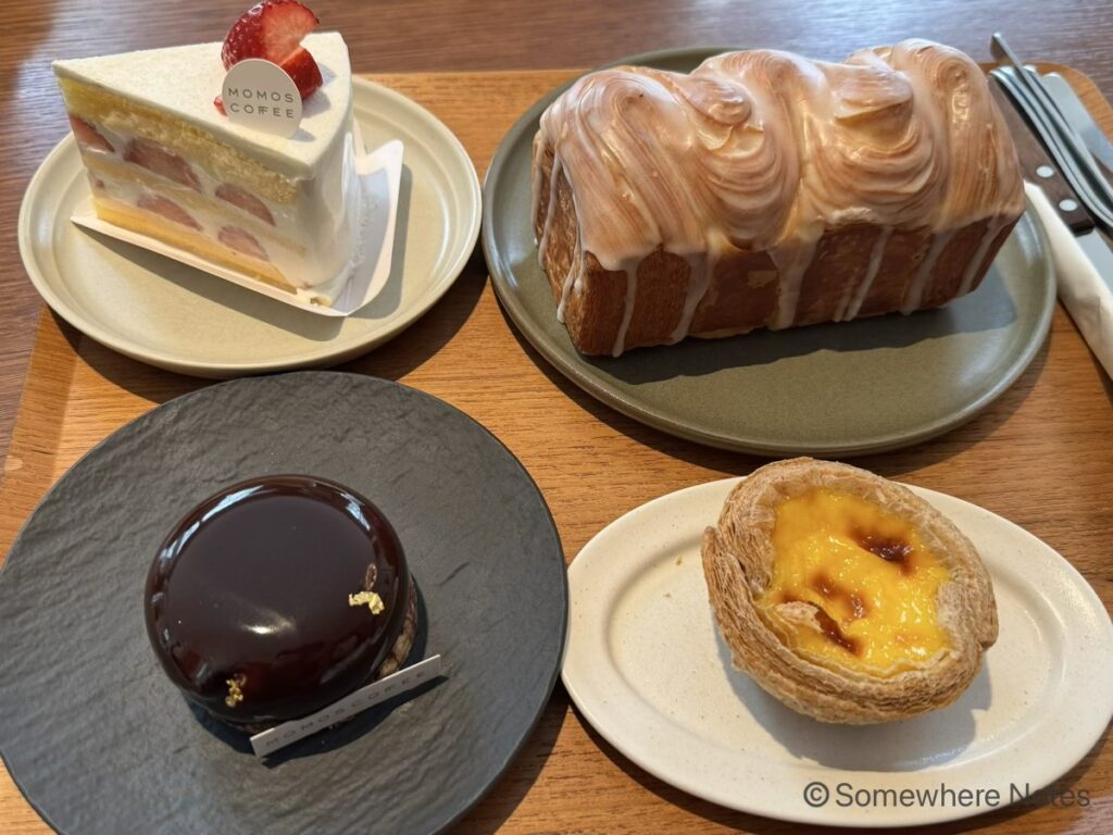 木製トレイに載った、釜山のモモスコーヒーで注文した4種類のスイーツとパン。左上にはトップにカットしたいちごが載り「MOMOS COFFEE」のピックが刺さったショートケーキ、右上には柚子のアイシングがかかった大きなデニッシュパン、左下には金箔が添えられた丸いつやのあるチョコレートケーキ、右下にはパイ生地のエッグタルト。それぞれ異なる皿に盛り付けられている。