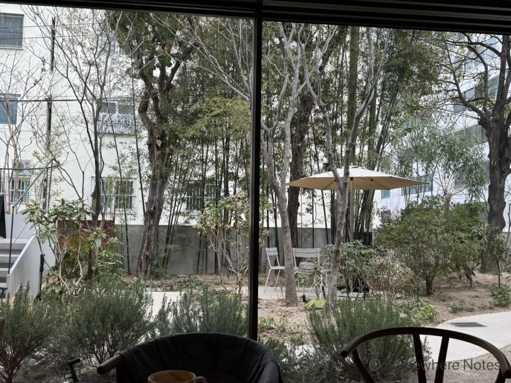 カフェの中から大きなガラス窓越しに見た、緑豊かな庭の風景。庭には竹林、木々、様々な植物が茂り、ベージュのパラソル、丸テーブル、白い椅子が置かれたテラス席がある。左側には外階段が見える。