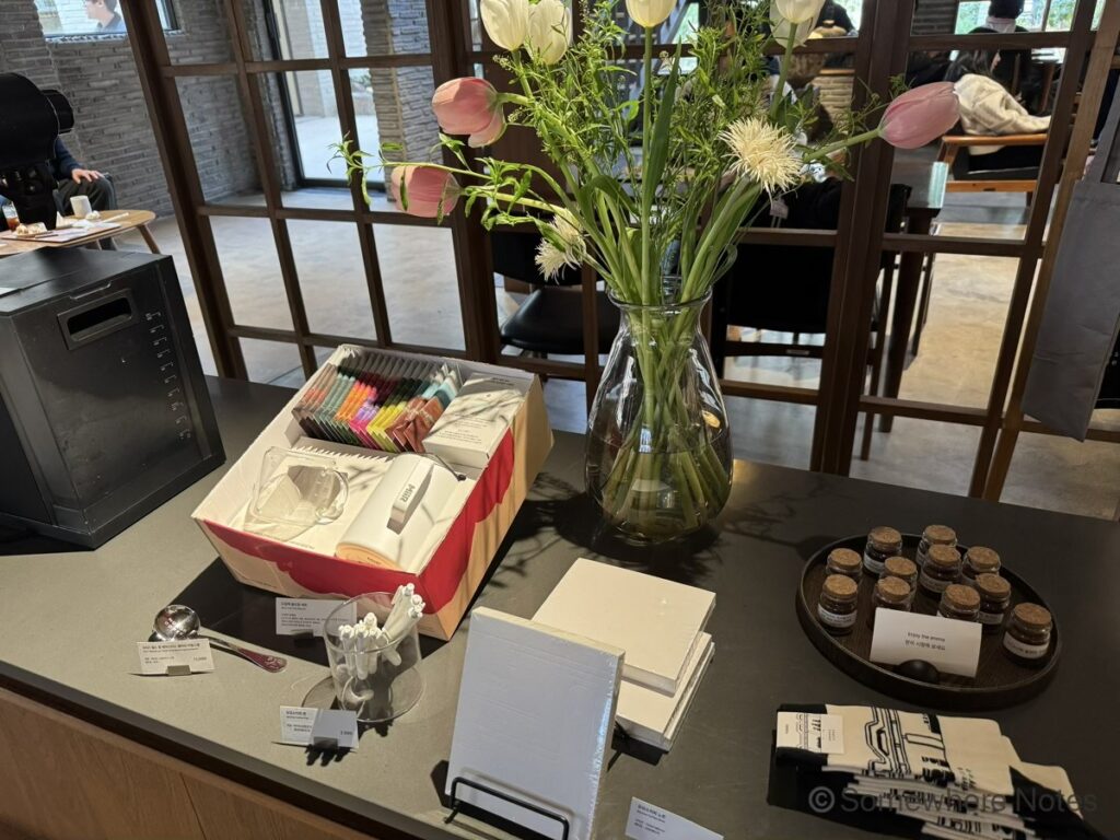 釜山のモモスコーヒー本店の店内カウンターに並ぶオリジナルグッズ。中央にはピンクのチューリップが飾られた大きなガラス花瓶。左側にはカラフルなドリップバッグやタンブラーが詰まったギフトボックスと白いペン。手前には無地のノート、右側にはコーヒーの香りを試せる小瓶が並ぶ丸いトレイや布製アイテム。背景には木枠のガラス窓とカフェでくつろぐ人々の姿。