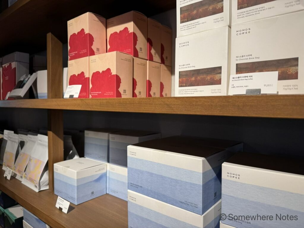 モモスコーヒー本店の木製陳列棚に並ぶ、お土産用のドリップコーヒー。上段には赤い花柄のパッケージや風景が描かれた箱入りのコーヒー。下段には水色の爽やかなデザインの大きなギフトボックスや袋入りのコーヒー豆。商品の手前にはハングル表記の値札。