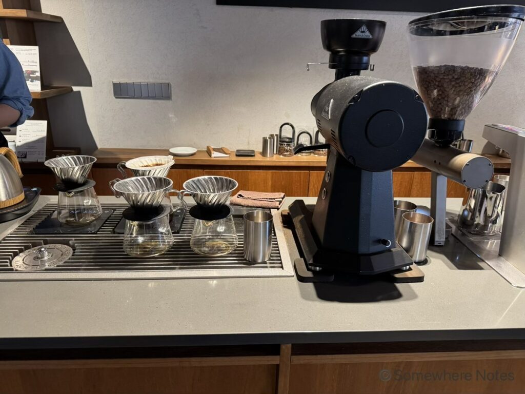 モモスコーヒー本店の店内カウンターの様子。左側にはハンドドリップ用のガラス製サーバーとドリッパーが金属製のトレイの上に並んでおり、右側には存在感のある大きな黒いコーヒー豆を挽く機械と、豆がたっぷり入った透明な容器が設置されている。奥の木製の棚にも器具が美しく整頓されており、プロフェッショナルな空間であることが伝わる。