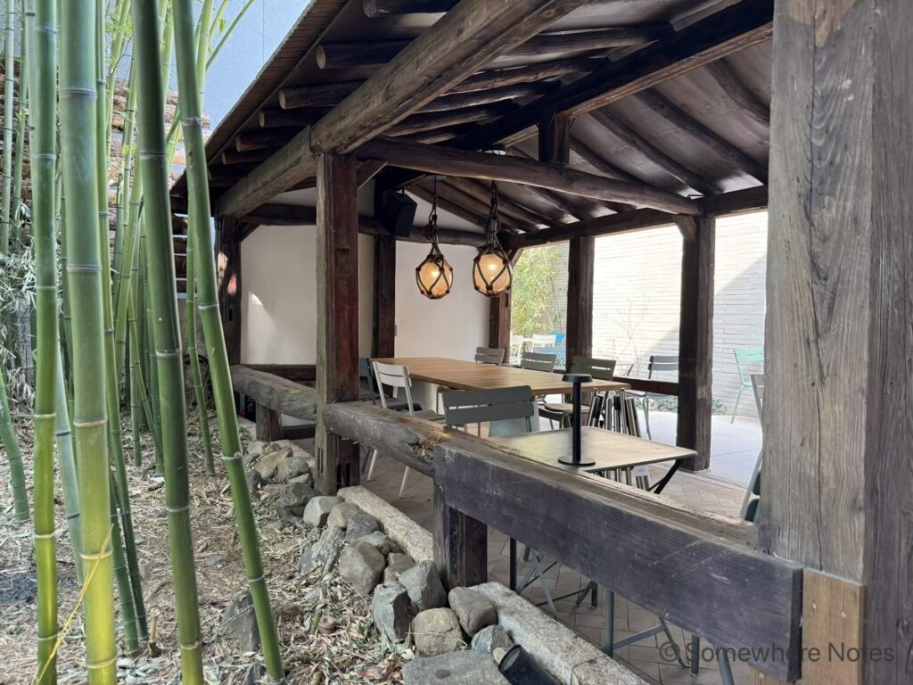 釜山のモモスコーヒー本店にある、太い木の柱と梁で組まれた屋根付きの屋外テラス席。天井から吊るされた、丸く温かい光を放つ2つのペンダントライト。中央には長方形の木製テーブルと複数の椅子。左側には青々とした竹林と足元に並ぶ自然石。