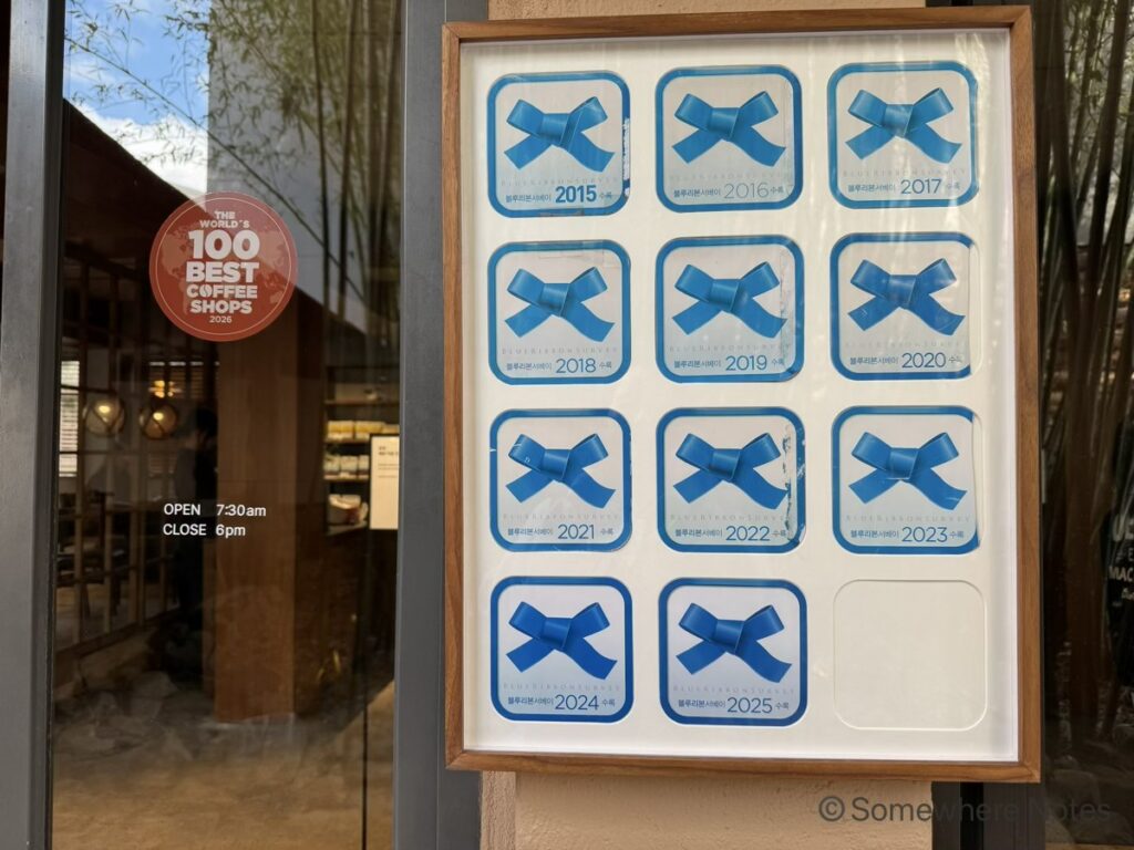 釜山のモモスコーヒー本店入り口。右側の壁には、韓国のグルメガイド「ブルーリボンサーベイ」に2015年から2025年まで連続選出されたことを示す、青いリボンのステッカーが並ぶ額縁。左側のガラスドアには「THE WORLD'S 100 BEST COFFEE SHOPS 2026」の赤い丸型ステッカーと営業時間。