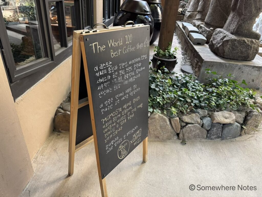釜山のモモスコーヒー本店の店先に置かれた、木枠の黒板の立て看板。上部にはチョークで「The World 100 Best Coffee Shops」と書かれ、その下にハングルで添えられた長文のメッセージ。背景にはカフェの建物の窓辺や緑の植物、大きな黒い壺、並んだ石像の足元。