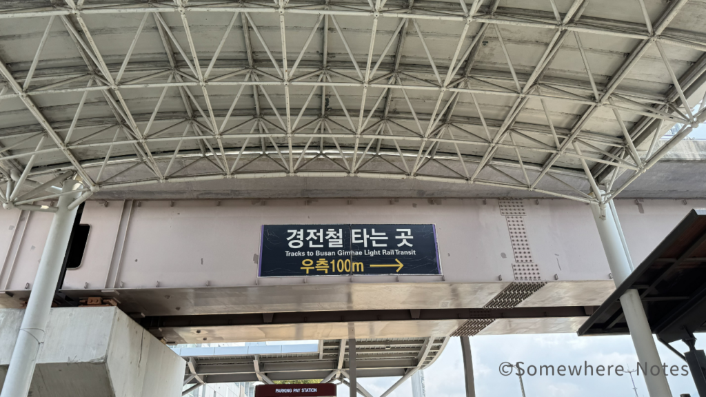 金海空港の屋外通路に「釜山金海軽電鉄」への案内板がある。軽電鉄乗り場、右へ100mと韓国語と記載あり。英語ではTracks to Busan Gimhae Light Rail Transitと書かれています。