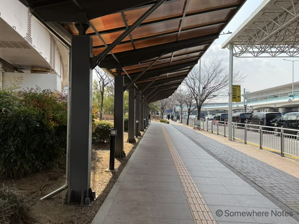 金海空港から軽電鉄「空港駅」へ向かう、屋根付きの歩道。左手に屋根が続き、足元には黄色の点字ブロックが真っ直ぐ伸びています。右奥に駅の建物へと続いています。