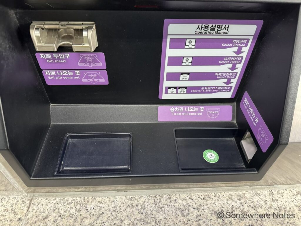 券売機の下部にある紙幣投入口と、切符・お釣りの取り出し口。右側の「切符が出る場所（Ticket will come out）」と書かれたトレイの中に、購入した緑色の丸いプラスチック製のトークンが一つ出てきている様子。