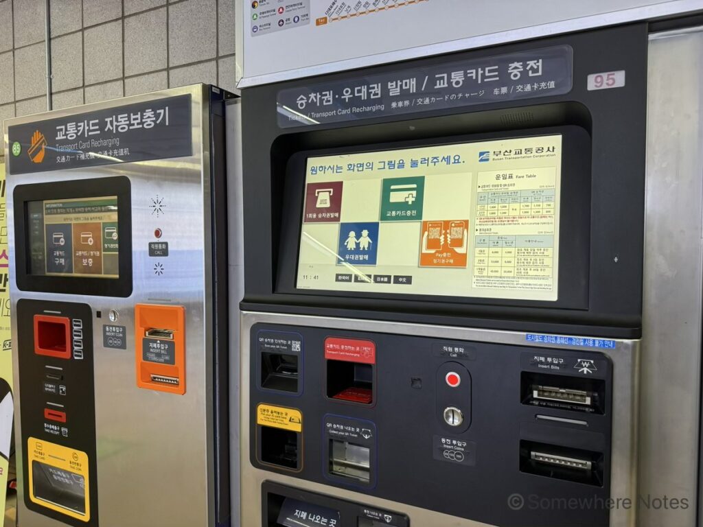 釜山地下鉄の自動券売機（右）と交通カードチャージ機（左）。券売機の画面には1回券 乗車券発売、交通カードチャージなどのメニューがイラスト付きで表示されており、画面下部のボタンで日本語表記への切り替えが可能です。