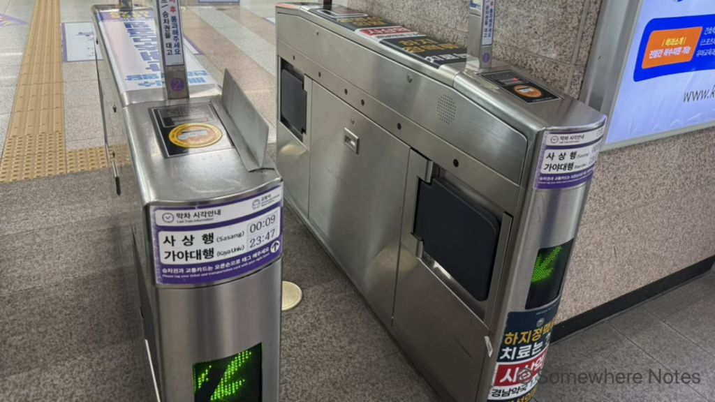 釜山金海軽電鉄の自動改札機。銀色の機体の上部には、交通カードやトークンをタッチする黄色の丸いセンサーがあり、手前側には通行可能を示す緑色の矢印が点灯しています。機体の側面にはササン行などの終電の時刻が書かれた紫色の案内が貼られています。